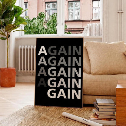 A black framed wall art print leaning against a beige sofa in a bright living room. The print features the text 'AGAIN AGAIN AGAIN AGAIN A G N' in large white and gray letters.