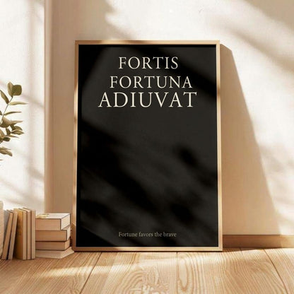 A framed black art print with the Latin phrase 'FORTIS FORTUNA ADIUVAT' in white serif text, leaning against a beige wall on a light wood floor next to stacked books and a plant.