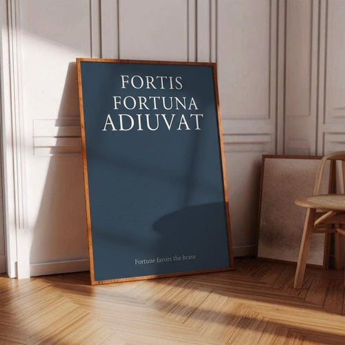 A framed art print with the Latin phrase 'FORTIS FORTUNA ADIUVAT' in white serif text on a dark blue background, leaning against a white paneled wall on a wooden floor in a sunlit room.