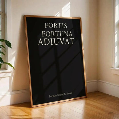 A framed black art print with the Latin text 'FORTIS FORTUNA ADIUVAT' and a smaller phrase near the bottom, leaning against a cream wall on a wooden floor in a sunlit room with windows and a plant.