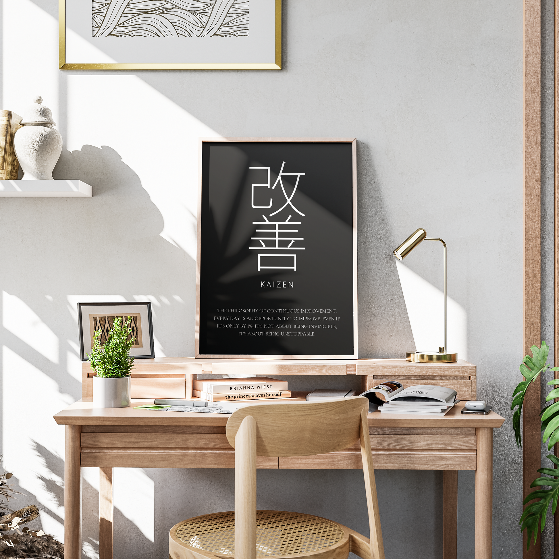 A framed black wall art print with large white Japanese characters and the word KAIZEN hangs above a light wood desk in a styled home office. The scene includes a wooden chair, gold desk lamp, books, plants, shelf decor, and warm natural light casting shadows on a gray wall.