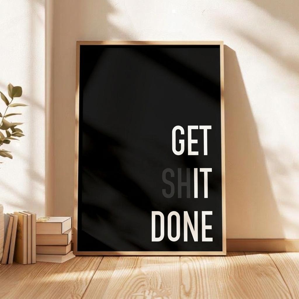 A framed motivational wall art print with the text 'GET SHIT DONE' in white on a black background, leaning against a beige wall on a wooden floor next to stacked books and a plant.