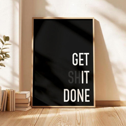 A framed motivational wall art print with the text 'GET SHIT DONE' in white on a black background, leaning against a beige wall on a wooden floor next to stacked books and a plant.