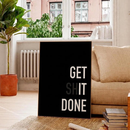 A black vertical wall art print with the motivational text 'GET SHIT DONE' in white and gray lettering, displayed in a cozy living room setting next to a beige sofa and a potted plant.