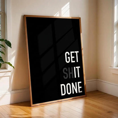 A framed black wall art print leaning against a beige wall on a wooden floor, with the text 'GET SHIT DONE' in white and gray. Sunlight streams in from two windows, and a green plant is visible at the left window.