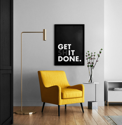 A framed black wall art print with the words 'GET IT DONE.' in white text hangs on a light gray wall above a mustard yellow armchair in a modern room with a gold floor lamp, wood floor, and a small side table with a vase of branches.