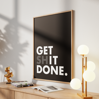 A framed wall art print with the text 'GET SHIT DONE.' in bold white letters on a black background, leaning on a light wood console table in a styled room with a lamp, books, and shadows on the wall.