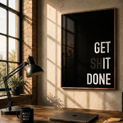 A framed black wall art print with bold white text reading 'GET SHIT DONE' hangs on a textured beige wall above a wooden desk in a styled office setting with a lamp, laptop, mug, and whiteboard.