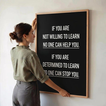 A woman in a sage green shirt and gray trousers stands beside a large framed motivational sign on a white wall. The sign has a black background with white text reading, 'IF YOU ARE NOT WILLING TO LEARN NO ONE CAN HELP YOU.
