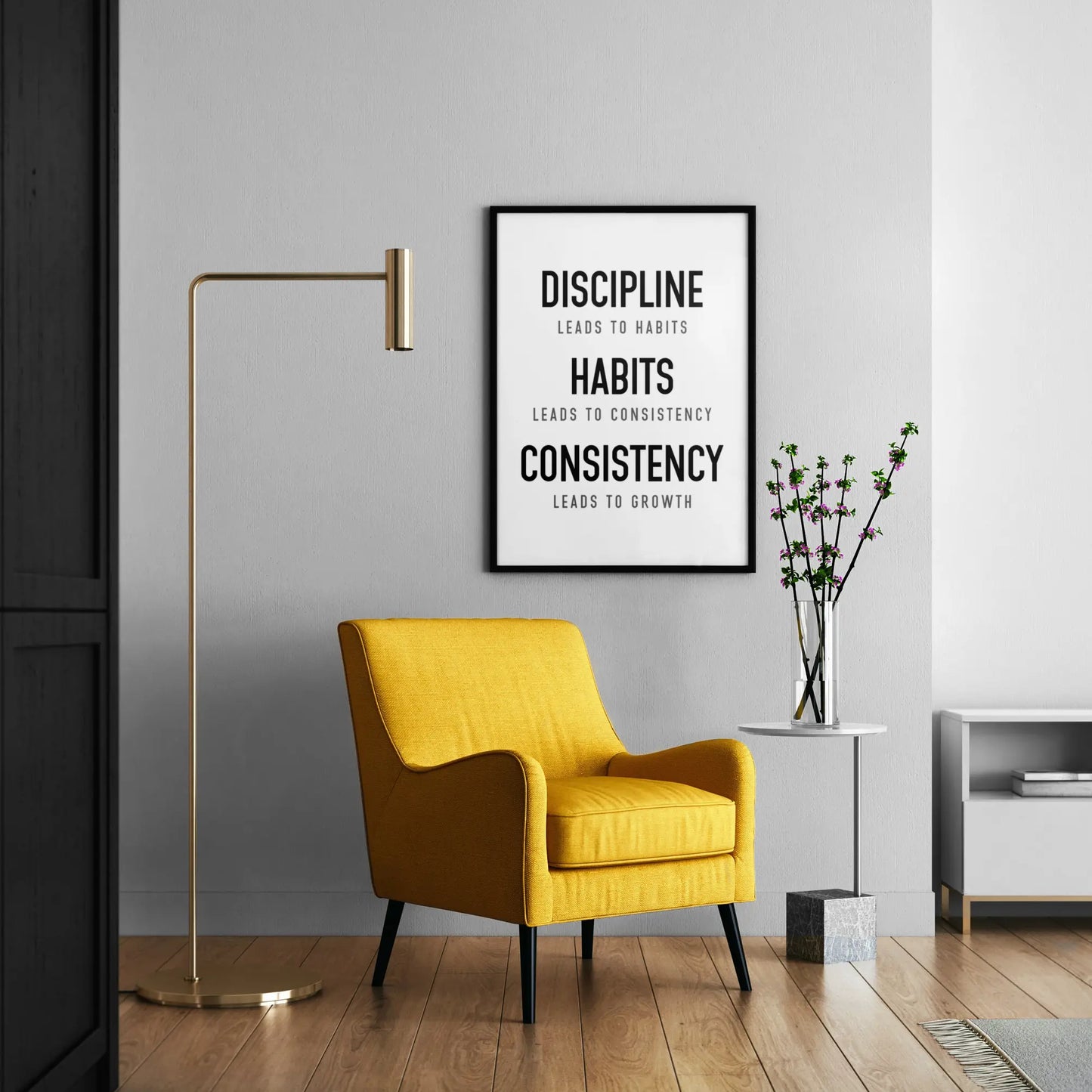 A framed motivational poster with black text on a white background hanging on a light gray wall in a modern living room setting with a yellow armchair, gold floor lamp, and side table.