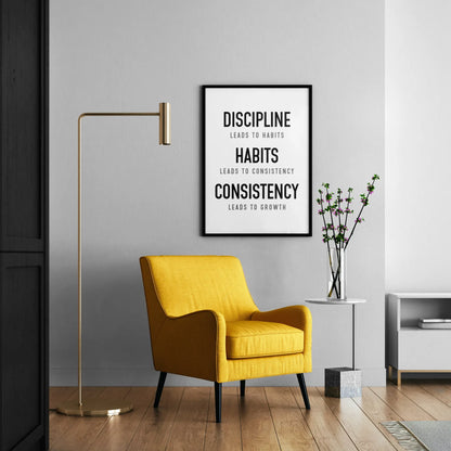A framed motivational poster with black text on a white background hanging on a light gray wall in a modern living room setting with a yellow armchair, gold floor lamp, and side table.