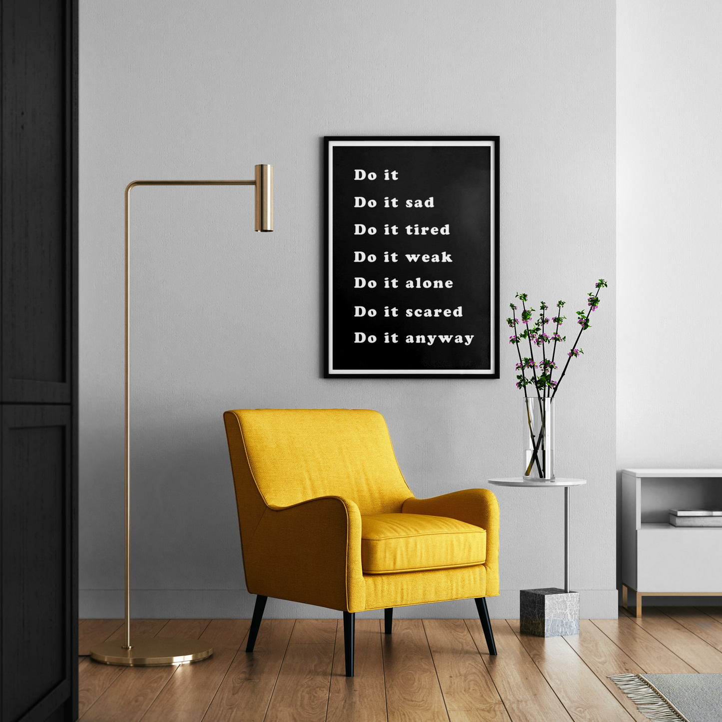 A framed black-and-white motivational poster with the text 'Do it sad, Do it tired, Do it weak, Do it alone, Do it scared, Do it anyway' hangs on a light gray wall above a mustard yellow armchair in a modern living room setting.