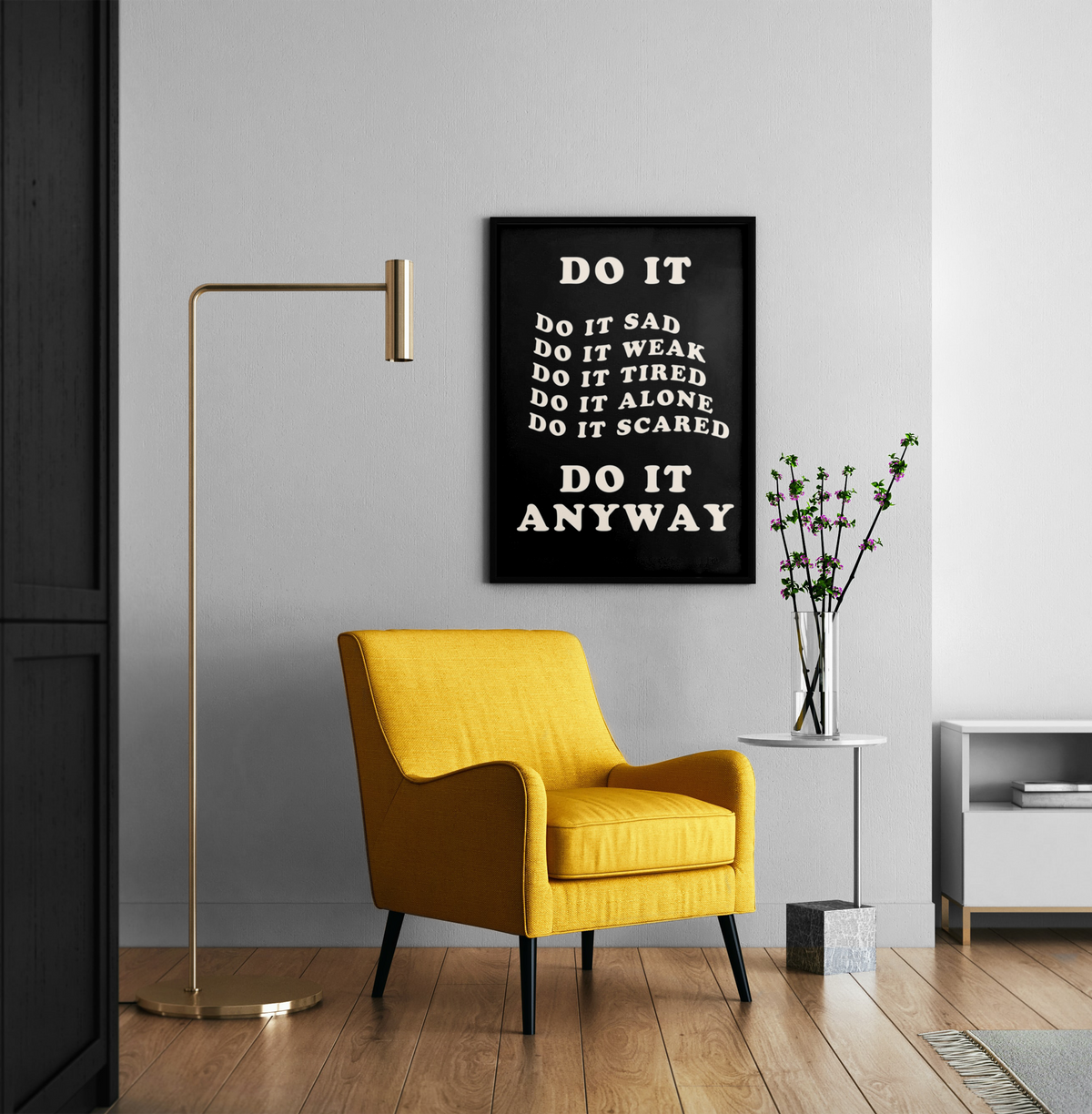 A black framed motivational poster with white text reading 'DO IT ANYWAY' hangs on a light gray wall in a modern living room setting with a yellow armchair, gold floor lamp, and side table.