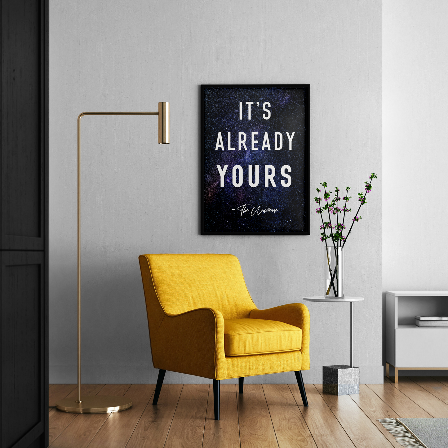 A framed wall art print with the quote 'IT'S ALREADY YOURS - To Ummah' hangs on a light gray wall in a modern living room setting with a yellow armchair, gold floor lamp, and side table.