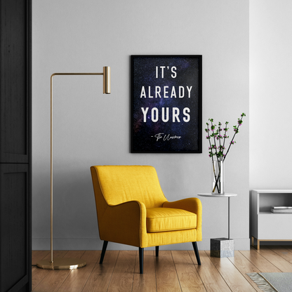 A framed wall art print with the quote 'IT'S ALREADY YOURS - To Ummah' hangs on a light gray wall in a modern living room setting with a yellow armchair, gold floor lamp, and side table.