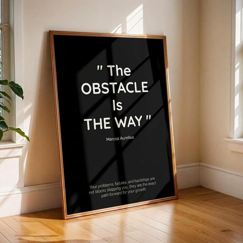 A framed black art print leaning against a beige wall on a wooden floor in a sunlit room. The print features a gold-toned frame and white text quoting Marcus Aurelius: "The Obstacle Is The Way" with additional motivational text below.