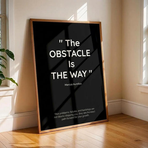 A framed black art print leaning against a beige wall on a wooden floor in a sunlit room. The print features a gold-toned frame and white text quoting Marcus Aurelius: "The Obstacle Is The Way" with additional motivational text below.