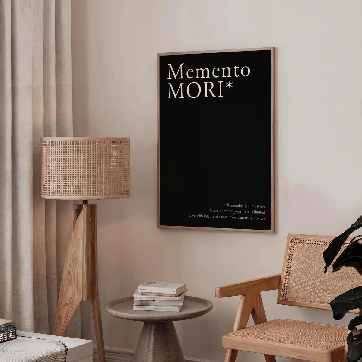 A framed black wall art print with the words 'Memento Mori' in white serif text hangs on a beige wall in a styled room with a floor lamp, small table, books, and a woven chair.