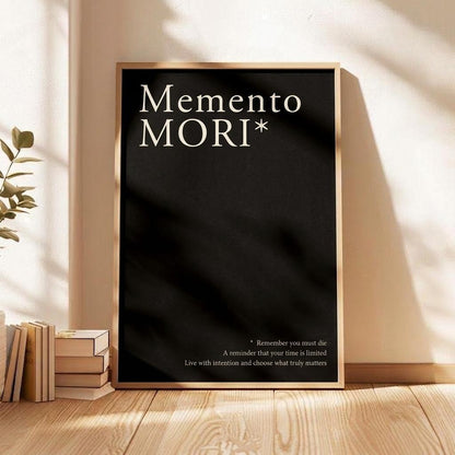 A framed black poster with the words 'Memento Mori*' in white serif text, leaning against a beige wall on a wooden floor beside stacked books and a plant.