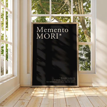 A framed black art print with the text 'Memento MORI*' in white serif lettering, leaning against a white wall in a bright room with wooden floors and windows showing outdoor greenery.