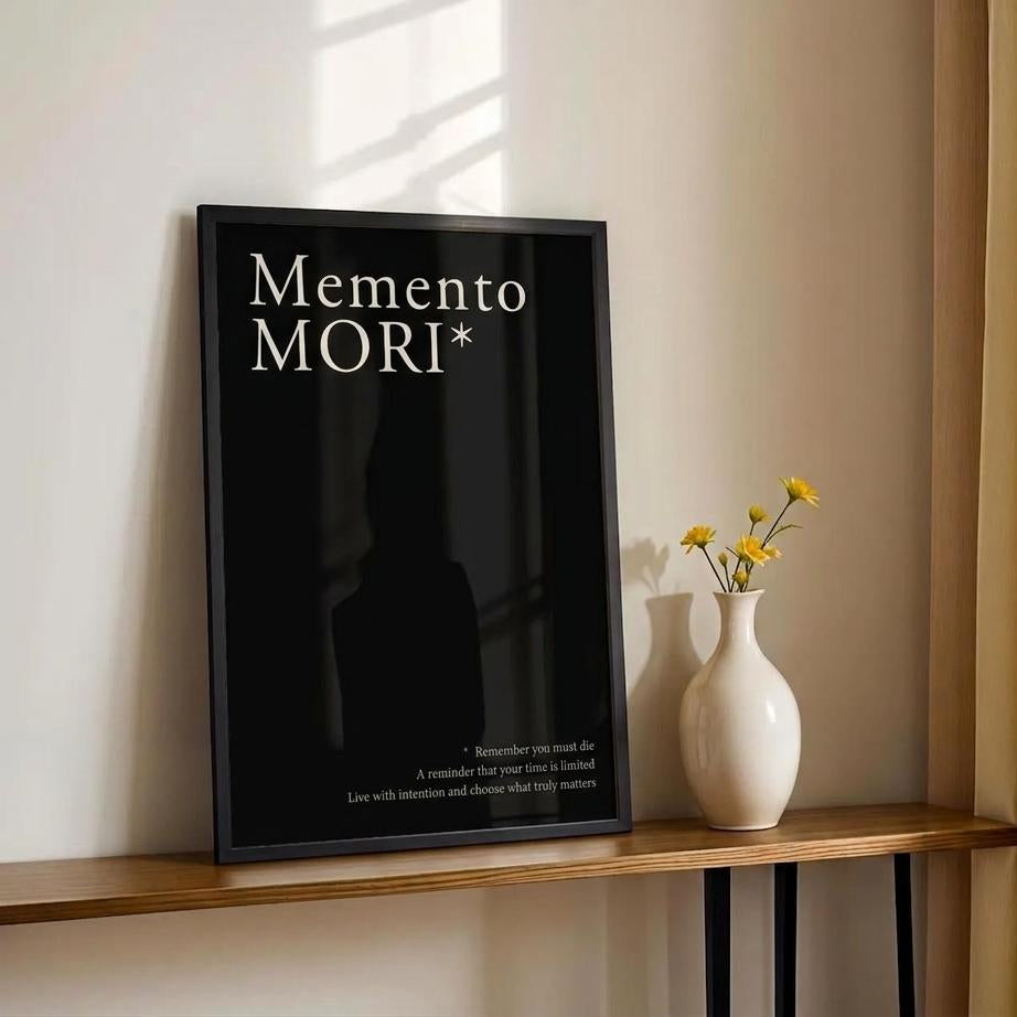A black framed wall art print with the words 'MEMENTO MORI' in white serif text, leaning on a wooden console table next to a white vase with yellow flowers against a beige wall and gold curtain.