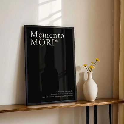 A black framed wall art print with the words 'MEMENTO MORI' in white serif text, leaning on a wooden console table next to a white vase with yellow flowers against a beige wall and gold curtain.