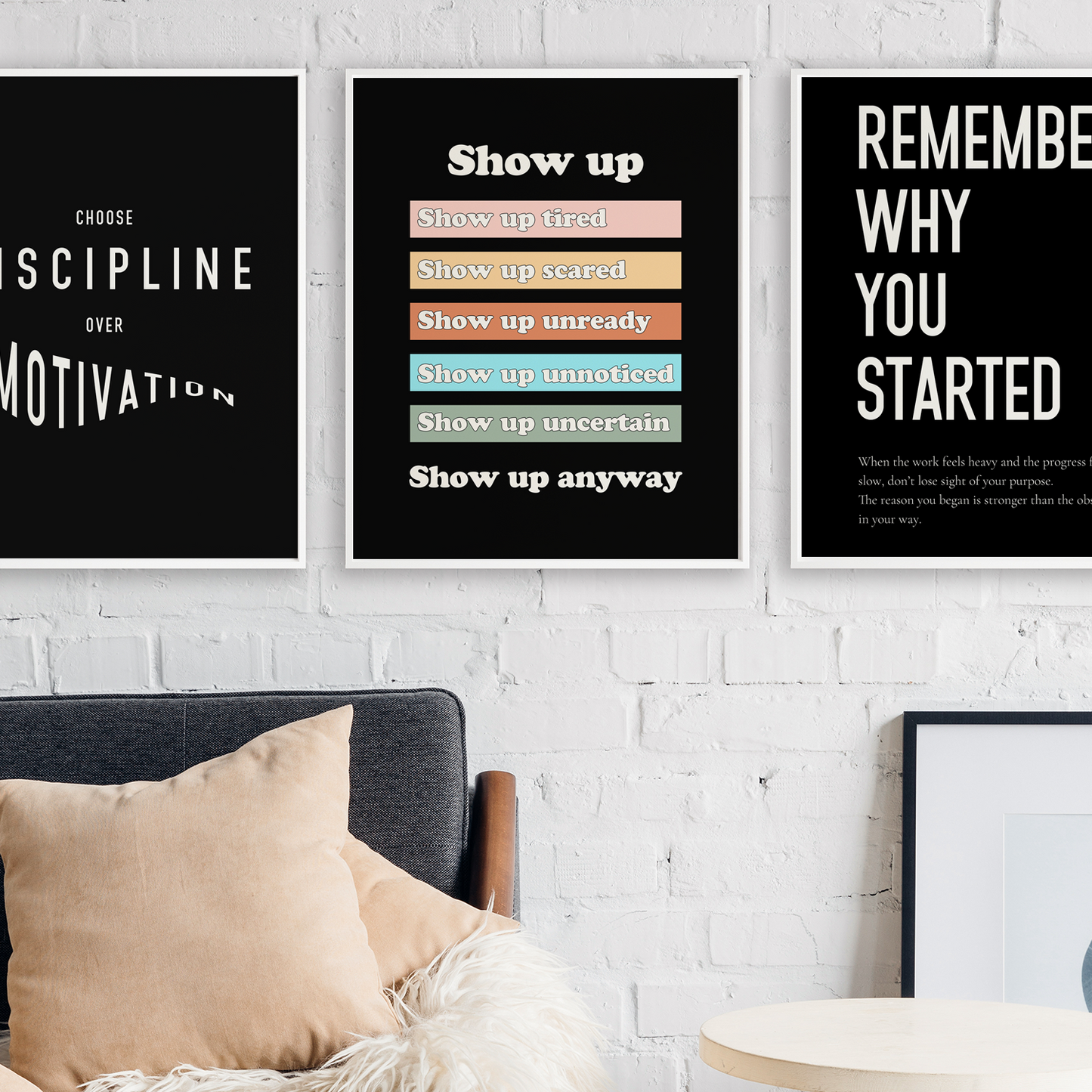Three framed motivational wall art prints with white text on black backgrounds hang on a white brick wall above a dark sofa with beige pillows. The center print has colored horizontal bars, and the right print includes a small quote at the bottom.