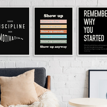 Three framed motivational wall art prints with white text on black backgrounds hang on a white brick wall above a dark sofa with beige pillows. The center print has colored horizontal bars, and the right print includes a small quote at the bottom.