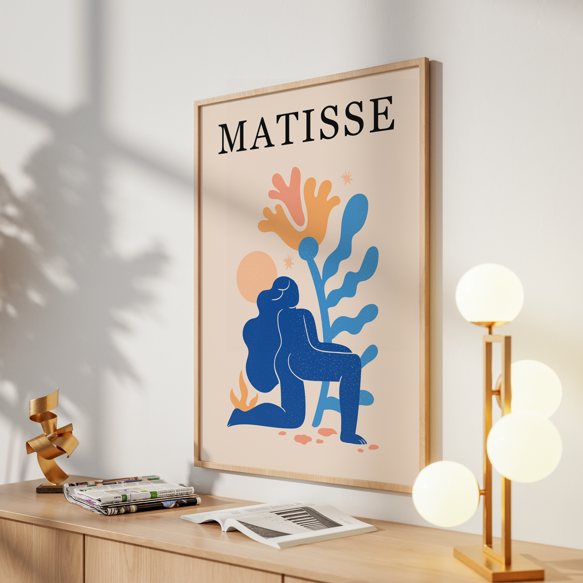 A framed art print featuring an abstract blue figure and coral-like shapes with the word 'MATISSE' at the top, displayed on a light wood console against a white wall with decorative objects around it.