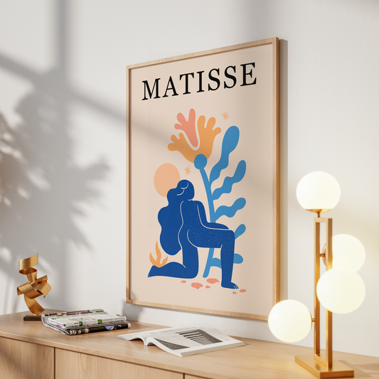 A framed art print featuring an abstract blue figure and coral-like shapes with the word 'MATISSE' at the top, displayed on a light wood console against a white wall with decorative objects around it.