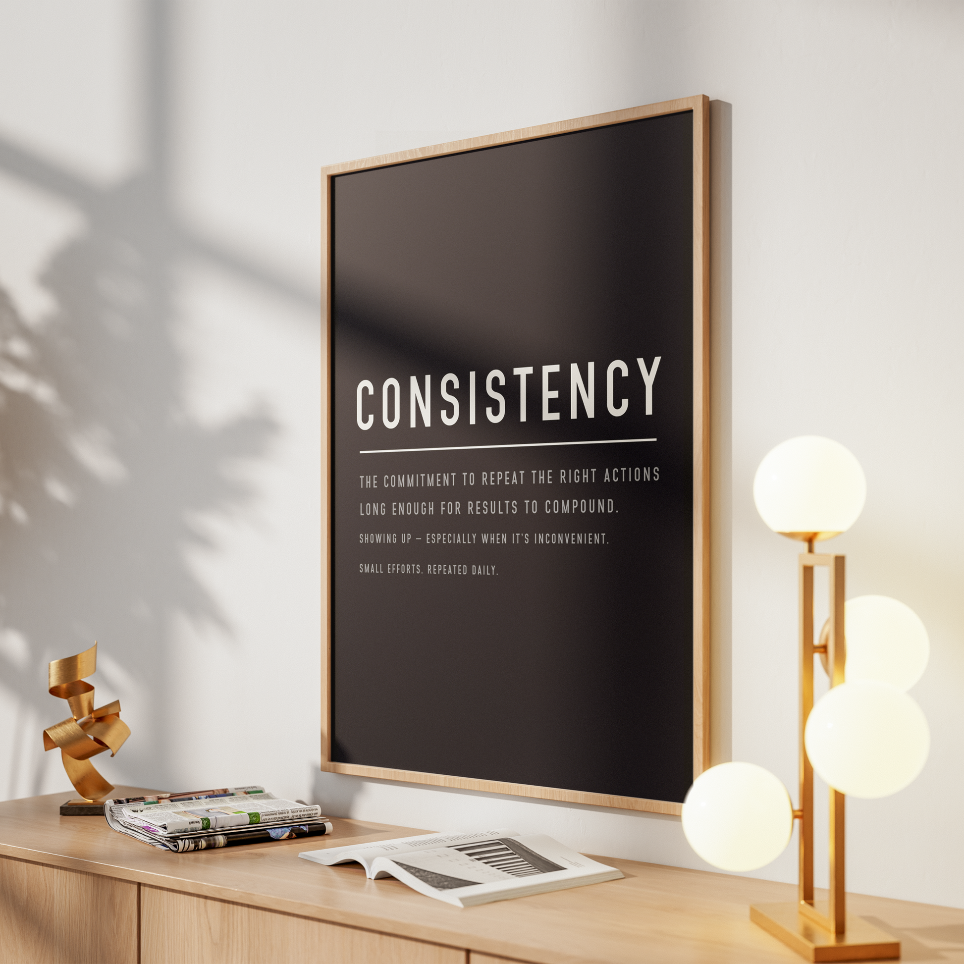 A framed wall art print with the word 'CONSISTENCY' in white text on a black background, leaning on a wooden console table in a modern room with a lamp, books, and shadows on the wall.