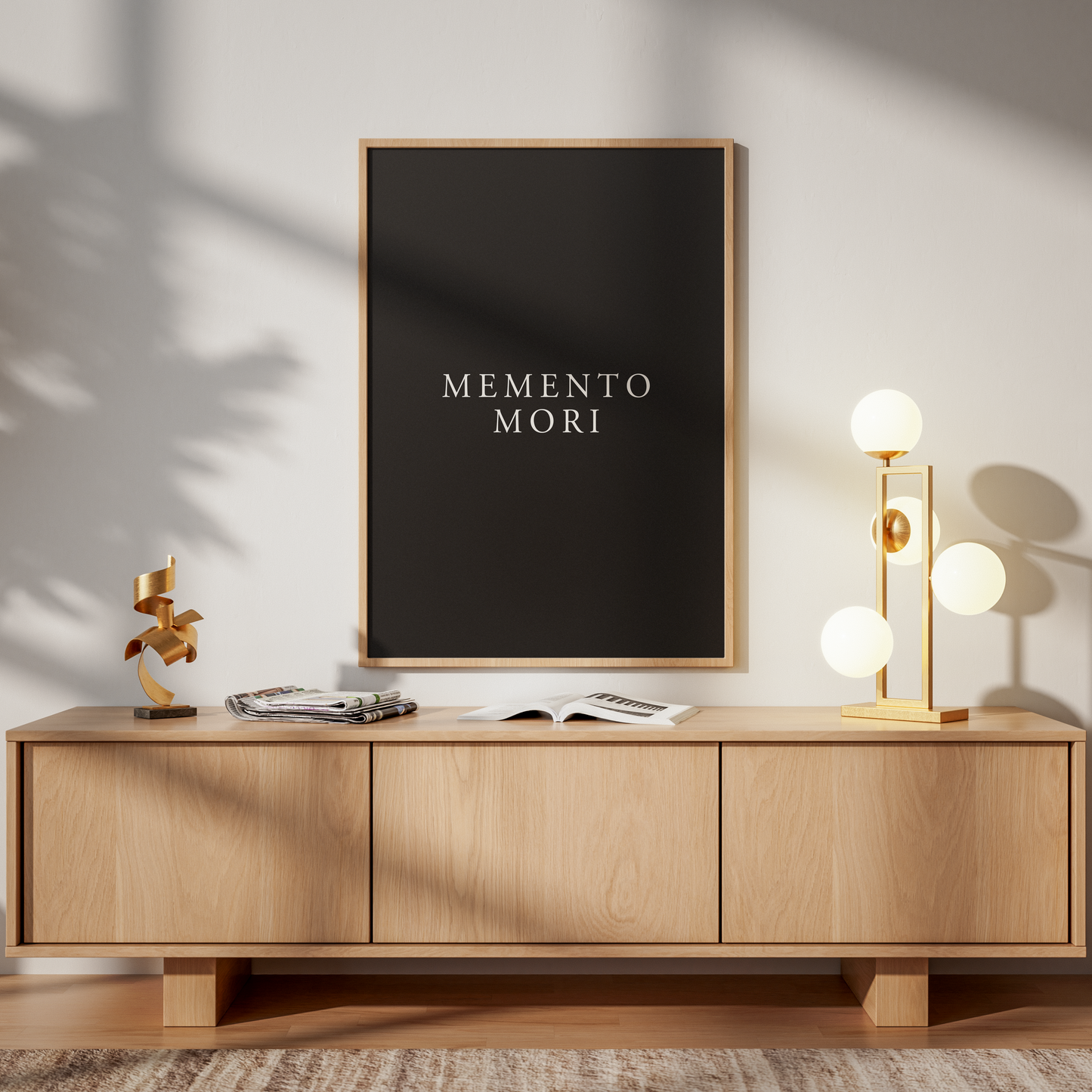 A framed black wall art print with the text 'MEMENTO MORI' in white, displayed above a light wood sideboard in a styled interior with warm sunlight and shadows.