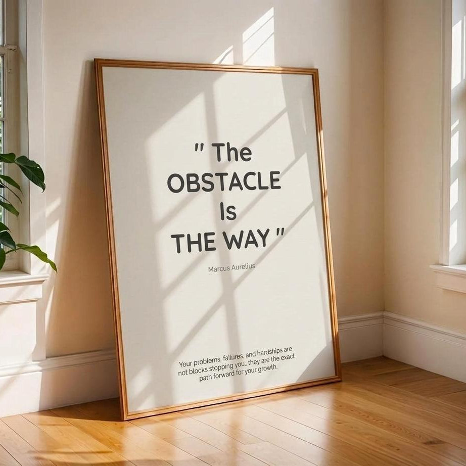 A framed motivational art print leaning against a beige wall on a wooden floor, featuring the quote 'The OBSTACLE IS THE WAY' by marcus aurelius. The print has a minimalist black text design on a white background with a light wood frame, set in a sunlit room with windows and a plant.