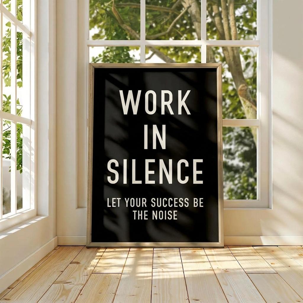 A framed black typographic wall art print with white text reading 'WORK IN SILENCE LET YOUR SUCCESS BE THE NOISE' leaning against a window in a bright room with wood floors and greenery visible outside.
