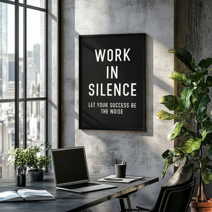 A black framed wall art print with white text reading 'WORK IN SILENCE LET YOUR SUCCESS BE THE NOISE' hanging on a textured gray wall in a modern office setting with a desk, laptop, and plants.