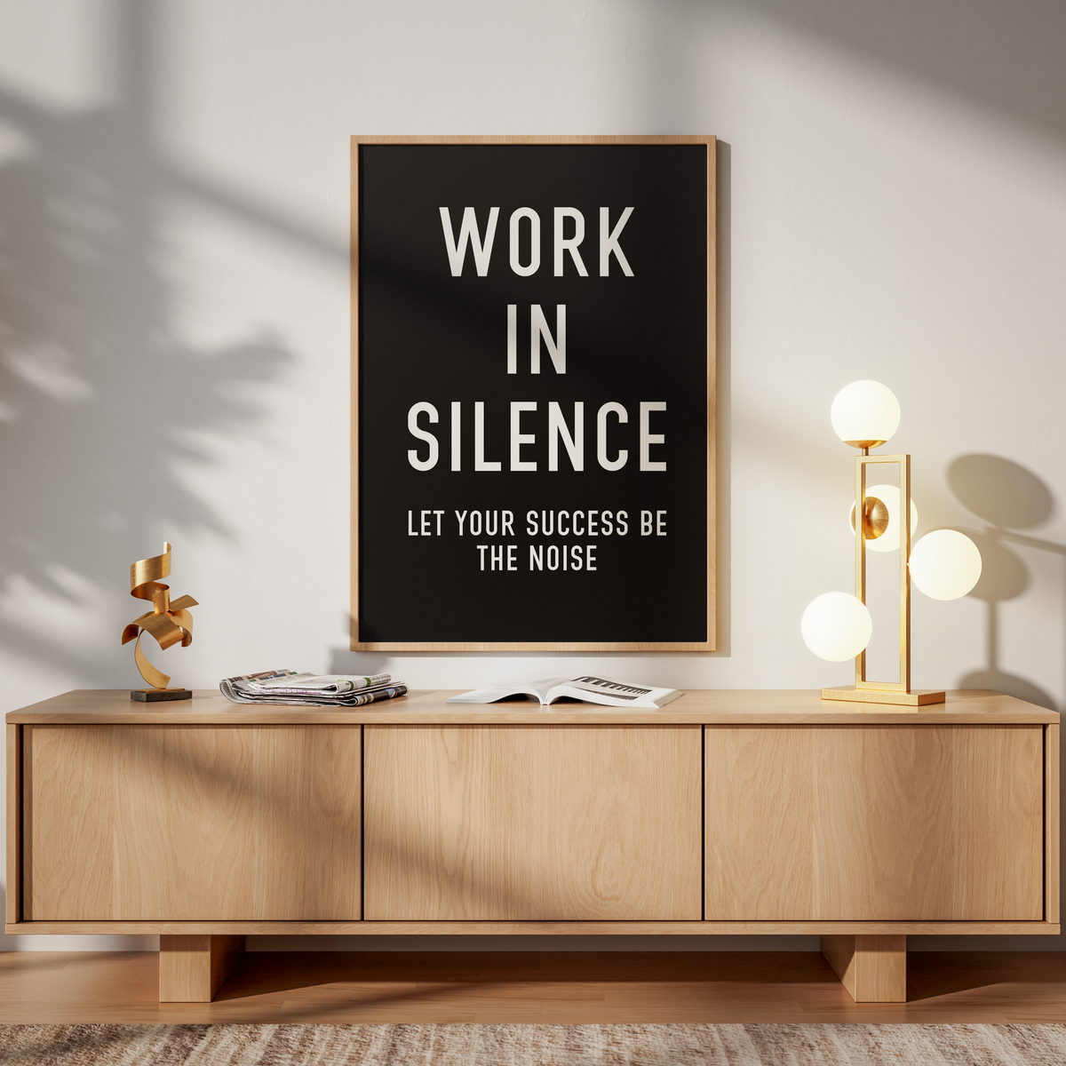A framed black poster with white text reading 'AMOR FATI CARPE DIEM' hangs above a light wood sideboard in a styled interior. The room has warm sunlight shadows, a gold floor lamp, books, and a wooden chair.