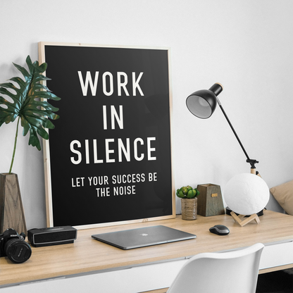 A framed black typographic wall art print with the words 'WORK IN SILENCE LET YOUR SUCCESS BE THE NOISE' in white lettering, displayed on a wooden desk in a modern home office setting with a lamp, laptop, and plant.