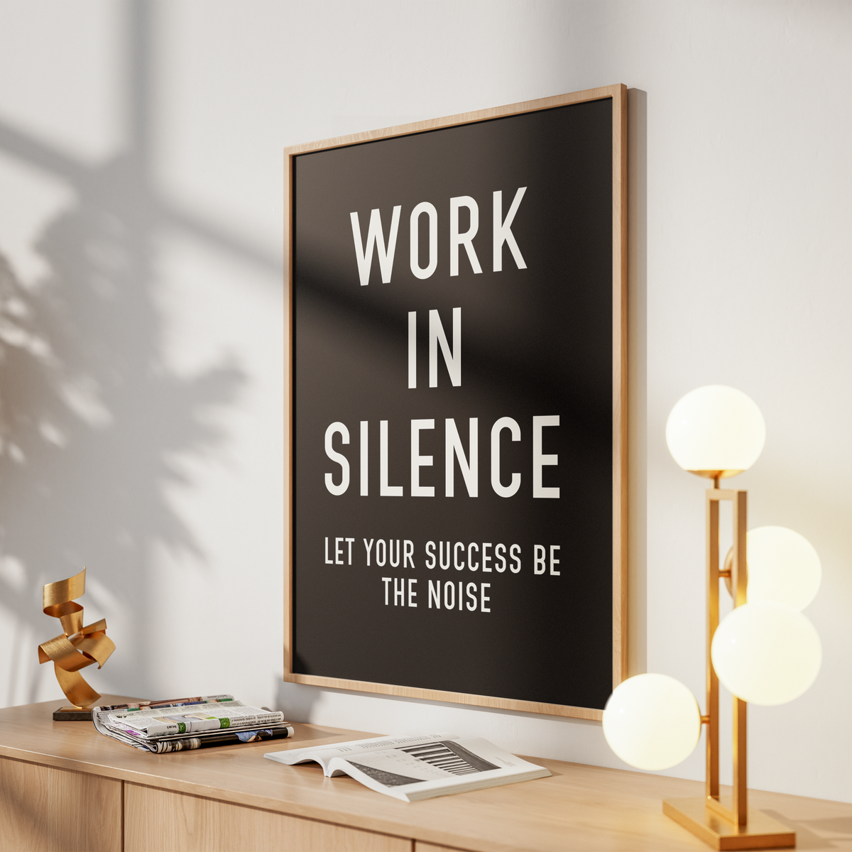 A framed wall art print with the text 'WORK IN SILENCE LET YOUR SUCCESS BE THE NOISE' in white on a black background, leaning on a wooden console in a styled room with a lamp, books, and a small sculpture.