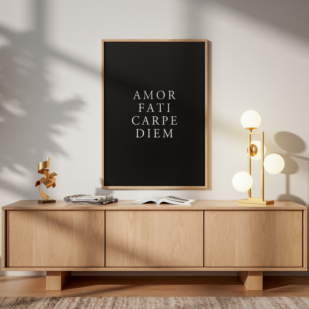 A framed black poster with white text reading 'AMOR FATI CARPE DIEM' hangs above a light wood sideboard in a styled interior. The room has warm sunlight shadows, a gold floor lamp, books, and a wooden chair.