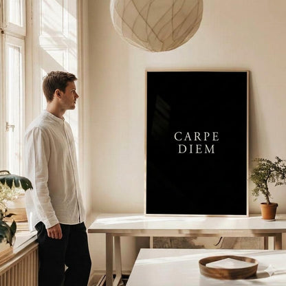 A man in a white shirt and black pants stands in a bright room looking at a large black framed wall art print with the text 'CARPE DIEM' in white serif letters.