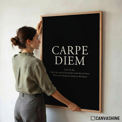 A woman in a sage green shirt and gray pants holds up a large framed wall art print with a black background and the words 'CARPE DIEM' in white, mounted on a light gray wall.