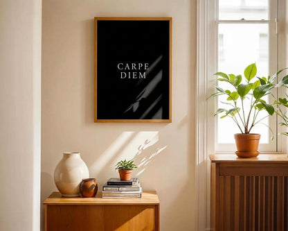 A framed black art print with the text 'MEMENTO MORI' in white serif letters, hanging on a beige wall in a sunlit room with a wooden side table, ceramic vases, and a potted plant by the window.