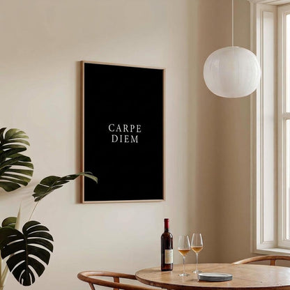A framed black wall art print with the words 'CARPE DIEM' in white serif text hangs on a beige wall in a bright dining room. A wooden table with two wine glasses and a bottle, a white pendant light, and a window are visible.