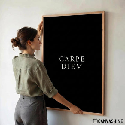 A woman in an olive green shirt and gray trousers is adjusting a large framed wall art print with the text 'CARPE DIEM' on a black background. The print has a light wood frame and is mounted on a white wall.
