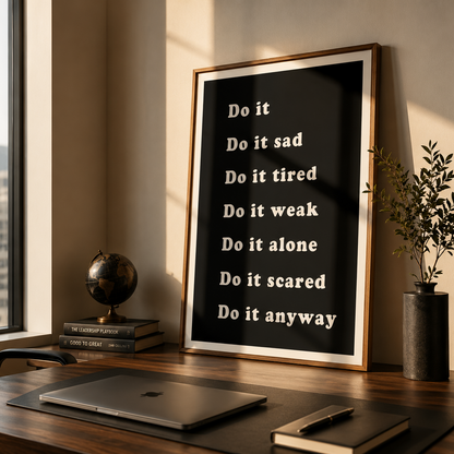 A framed motivational wall art print with white text on a black background, leaning on a wooden desk in a modern office setting with a laptop, books, and a window view.