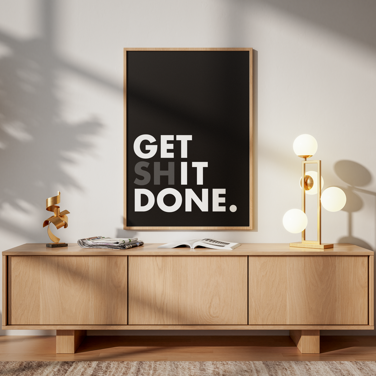 A framed wall art print with the text 'GET SHIT DONE.' in white on a black background, displayed above a light wood sideboard in a styled room with a lamp, chair, and plant shadows.