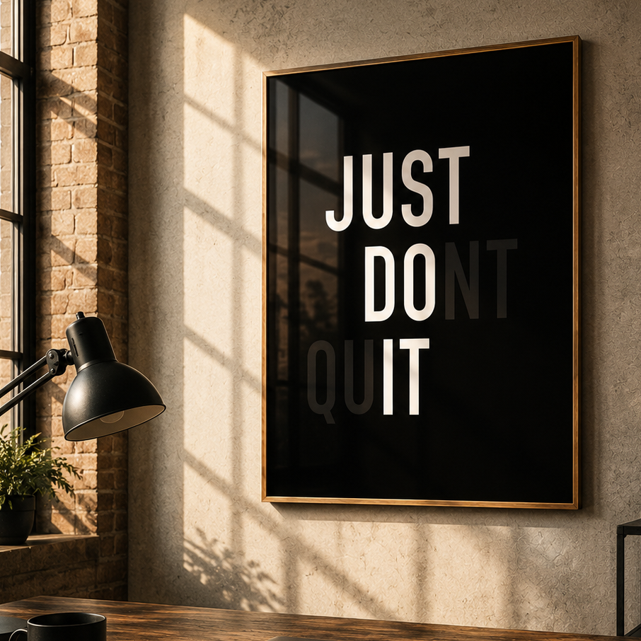 A framed motivational wall art print with the text 'JUST DO IT' in bold white letters on a black background, hanging on a beige wall in a styled home office setting with a desk, laptop, lamp, and whiteboard.
