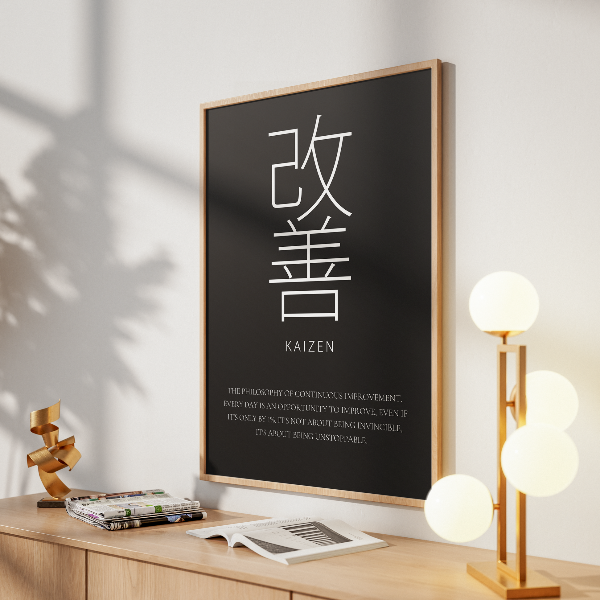 A framed wall art print with a black background and large white Japanese characters, plus the word 'KAIZEN' and a short English quote, displayed above a wooden console table in a styled interior.