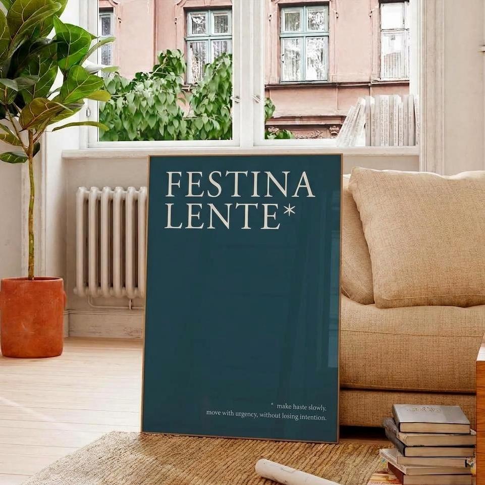 A large dark teal art print or book cover with the words 'FESTINA LENTE*' in white serif lettering, standing on a woven rug in a bright living room beside a beige sofa, a potted plant, and a stack of books.