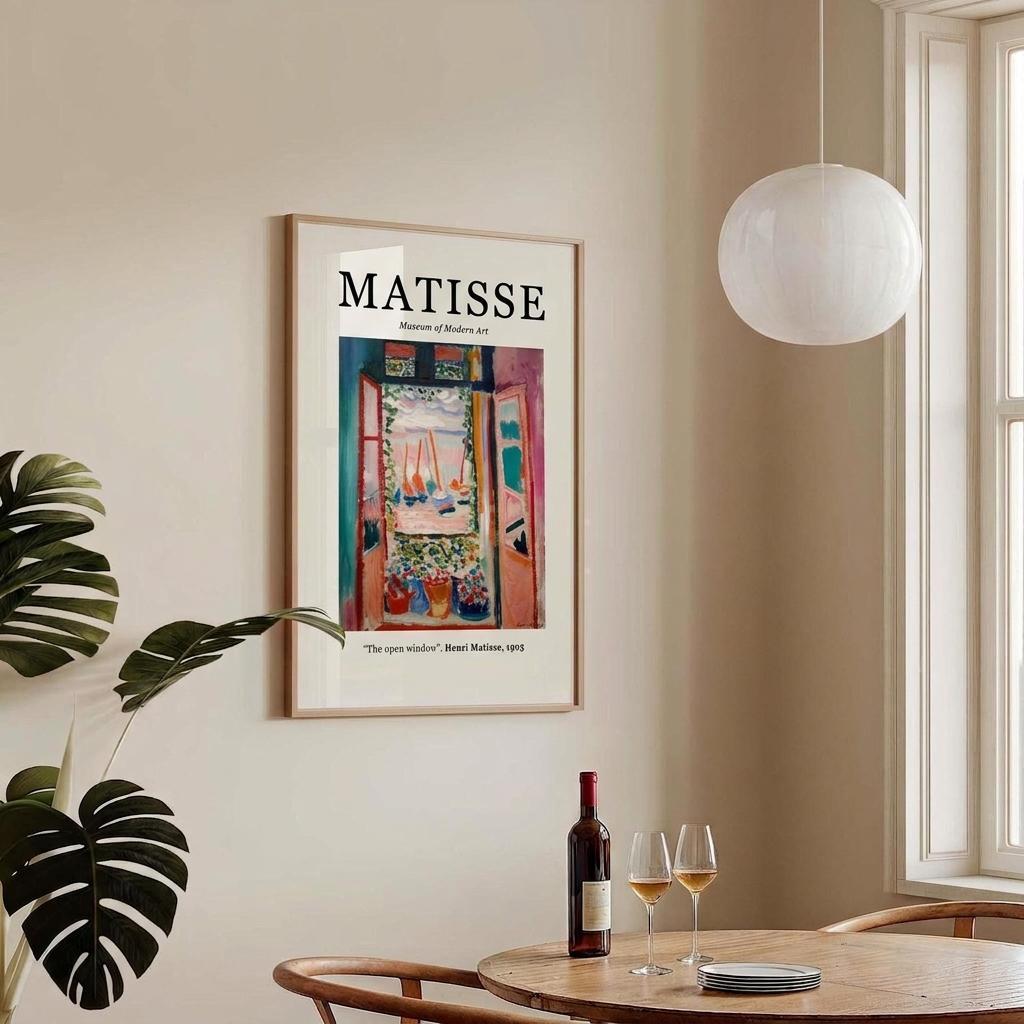 A framed art print of Henri Lebasque's Matisse 'The Open Window' poster hanging on a beige wall in a modern dining room setting with a wooden table, wine bottle, and glasses, alongside a white globe pendant light and a green monstera plant.
